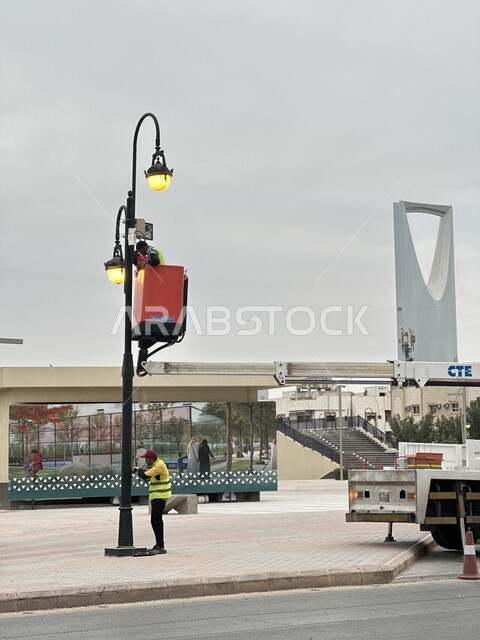 تحسين الخدمات وتعزيز الأمان في السعودية، عمال عربيون خليجيون سعوديون يستخدمون رافعة مثبتة على شاحنة للوصول إلى أعلى العامود، تركيب كاميرات مراقبة في جميع مدينة أنحاء الرياض، مراقبة المرور وضبط المخالفات والسرعة الزائدة، خلفية برج الممكلة المشهور في العاصمة