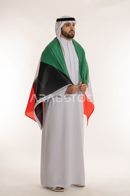 Standing straight and looking at something, love of country and belonging to it, showing feelings of love for the country, portrait of a young Kuwaiti Gulf Arab man wearing the agal, ghutra and Kuwaiti flag with pride and honor, national occasions and holidays, Kuwait National Day on February 25, full body image, white background