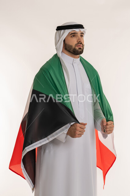 النظر إلى شيء ما واظهار مشاعر الحب والانتماء للبلاد, بورتريه لشاب عربي خليجي كويتي يرتدي الثوب التقليدي والغترة والعلم الكويتي بفخر واعتزاز, مناسبات وأعياد وطنية, اليوم الوطني لدولة الكويت في 25 فبراير، خلفية بيضاء