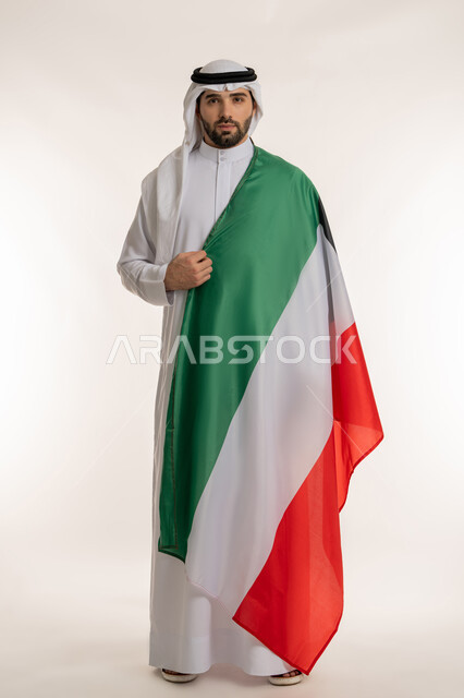 المناسبات والأعياد الوطنية, اظهار مشاعر الفخر بالانتماء للوطن, بورتريه لشاب عربي خليجي كويتي يرتدي الثوب التقليدي والغترة يضع على كتفه علم الدولة وينظر إلى الكاميرا باعتزاز, استذكار اليوم الوطني الكويتي في 25 فبراير, صورة الجسم بالطول الكامل, خلفية بيضاء