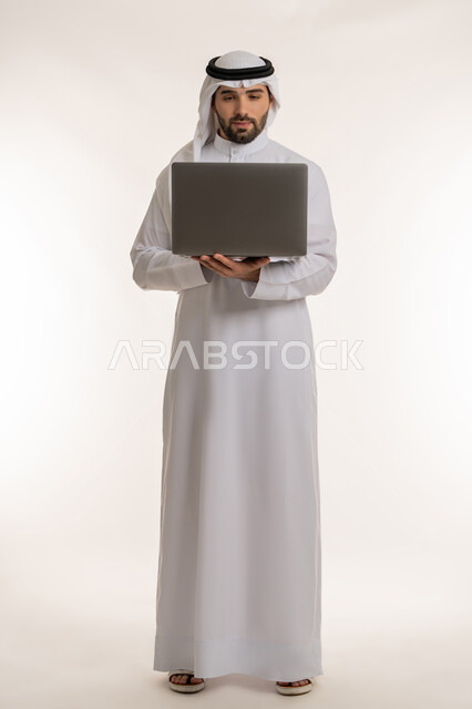 Standing up straight and completing tasks online, remote work concept, portrait of a young Saudi Arabian Gulf man wearing traditional thobe and ghutra using laptop, integrating work with technology and modern and advanced technology, full body image, white background