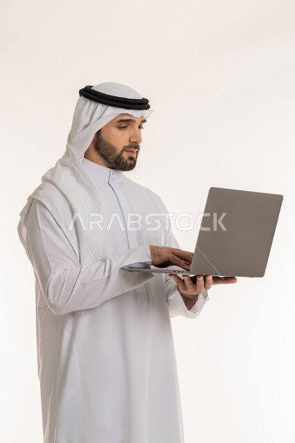 دمج العمل مع التقنية والتكنولوجيا الحديثة والمتطورة, انجاز وادارة المهام عبر الأونلاين, مفهوم العمل عن بعد, بورتريه لشاب عربي خليجي سعودي يرتدي الثوب التقليدي والغترة يستخدم الحاسب المحمول, التواصل والدردشة مع الأصدقاء والأهل عبر السوشال ميديا, خلفية بيضاء