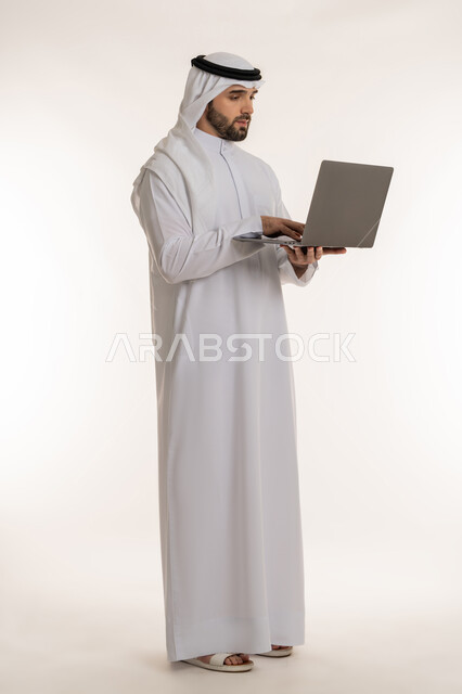 انجاز وادارة المهام عبر الأونلاين, مفهوم العمل عن بعد, بورتريه لشاب عربي خليجي سعودي يرتدي الثوب التقليدي والغترة يستخدم الحاسب المحمول, دمج العمل مع التقنية والتكنولوجيا الحديثة والمتطورة, صورة الجسم بالطول الكامل, خلفية بيضاء