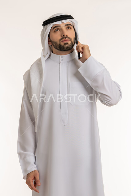 النظر إلى مكان ما ومحاولة تذكر شيء معين, الدردشة والتواصل مع الأهل والأصدقاء عبر السوشال ميديا, , بورتريه لشاب عربي خليجي سعودي يرتدي الثوب التقليدي والغترة يقوم بإجراء مكالمة صوتية عبر الهاتف المحمول, استخدام الاجهزة والتقنيات الحديثة والمتطورة، خلفية بيضاء