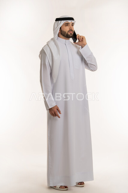 اجراء مكالمة صوتية عبر الهاتف المحمول, بورتريه لشاب عربي خليجي سعودي يرتدي الثوب التقليدي والغترة يمسك بيده الجوال, استخدام الاجهزة والتقنيات الحديثة والمتطورة, الدردشة والتواصل مع الأهل والأصدقاء عبر السوشال ميديا، صورة الجسم بالطول الكامل, خلفية بيضاء