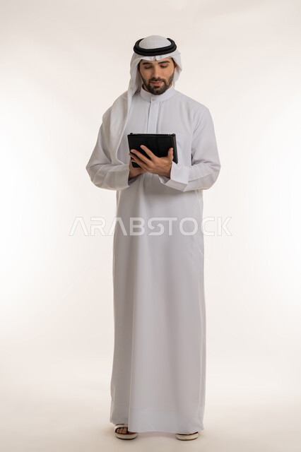 التواصل والدردشة مع الأهل والأصدقاء عبر السوشال ميديا, انجاز المهام عبر الأجهزة والتقنيات الحديثة والمتطورة, بورتريه لرجل عربي خليجي سعودي يرتدي الثوب التقليدي والغترة يحمل بيده الجهاز اللوحي, صورة الجسم بالطول الكامل, خلفية بيضاء