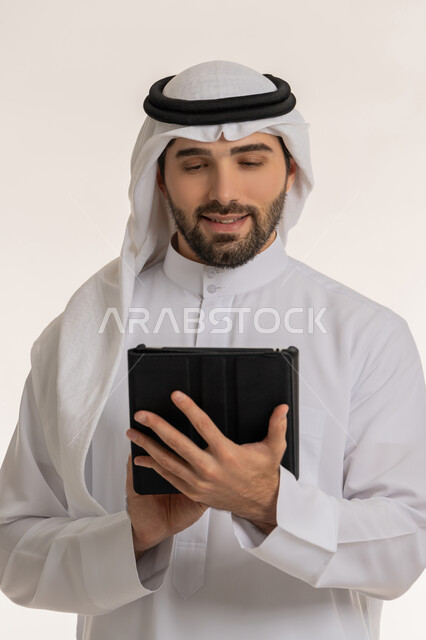 انجاز المهام عبر الأجهزة والتقنيات الحديثة والمتطورة, بورتريه مقرب لرجل عربي خليجي سعودي يرتدي الثوب التقليدي والغترة يحمل بيده الجهاز اللوحي, التواصل والدردشات مع الأهل والأصدقاء عبر السوشال ميديا, خلفية بيضاء