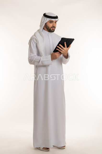 Communicating with friends via social media, remote business management, portrait of a young Saudi Arabian Gulf man wearing traditional thobe and ghutra holding a tablet, using modern and advanced devices and technologies, full-length body image, white background