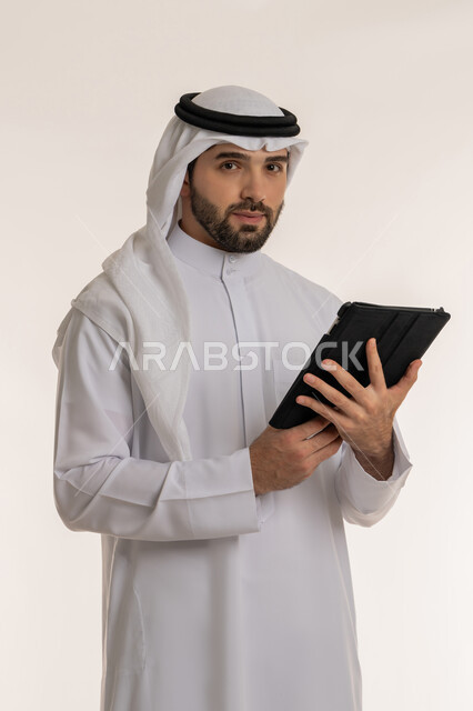 استخدام الاجهزة والتقنيات الحديثة والمتطورة, إدارة الأعمال عن بعد, التواصل مع الأصدقاء عبر وسائل التواصل الاجتماعي, بورتريه لشاب عربي خليجي سعودي يرتدي الثوب التقليدي والغترة ينظر إلى الكاميرا ويحمل بيده الجهاز اللوحي, خلفية بيضاء