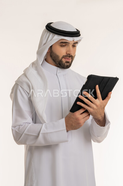Remote business management, communicating with friends via social media, portrait of a young Saudi Arabian Gulf man wearing traditional thobe and ghutra holding a tablet, using modern and advanced devices and technologies, white background