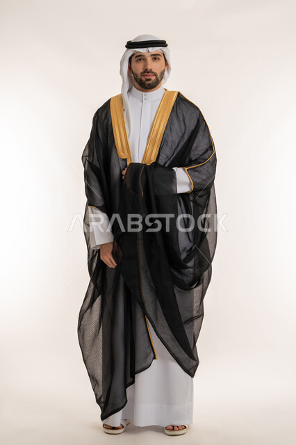 الوقوف باستقامة بإيماءات الثقة, مفهوم الاناقة والاهتمام بالمظهر الخارجي, بورتريه لشاب عربي خليجي سعودي يرتدي الثوب التقليدي والبشت ينظر إلى الكاميرا, صورة الجسم بالطول الكامل, خلفية بيضاء