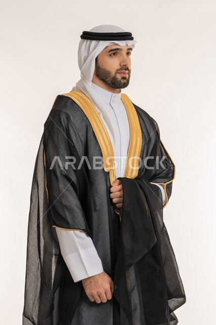 مفهوم الرجولة والاهتمام بالمظهر الخارجي, أناقة الشباب الخليجين, بورتريه لشاب عربي خليجي سعودي يرتدي الثوب التقليدي والبشت ينظر إلى شيء ما, الوقوف باستقامة وثقة بالنفس, خلفية بيضاء