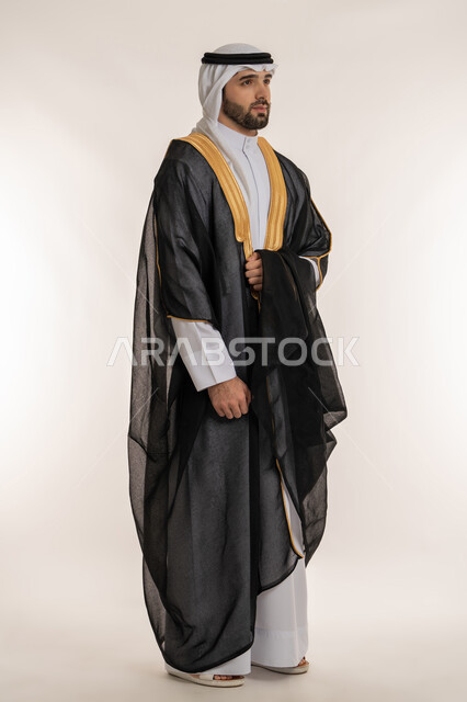 The concept of elegance and interest in external appearance, portrait of a young Saudi Arabian Gulf man wearing traditional dress and bisht looking at something, standing straight and confident, full body image, white background