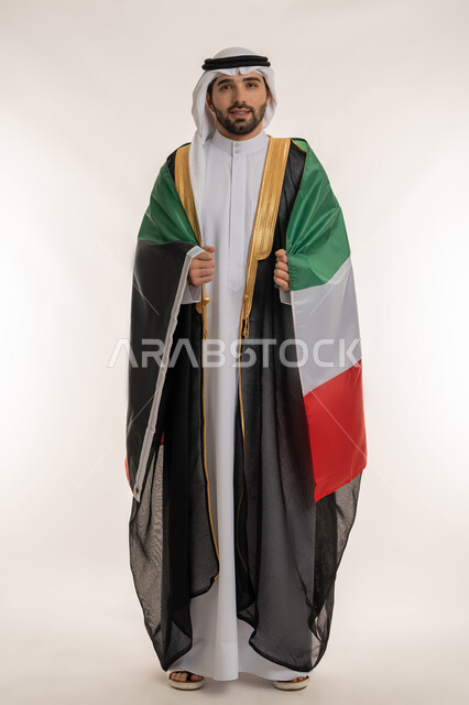 مناسبات وأعياد وطنية, اظهار مشاعر الفخر بالانتماء للوطن, بورتريه لشاب عربي خليجي كويتي يرتدي الدشداشة الكويتية والبشت يضع على كتفه علم دولة الكويت باعتزاز, استذكار اليوم الوطني الكويتي في 25 فبراير, صورة الجسم بالطول الكامل, خلفية بيضاء