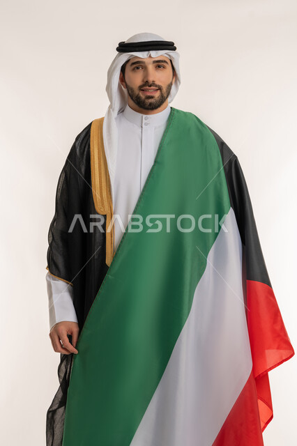 النظر إلى الكاميرا بايماءات الاعتزاز والفخر بإنجازات البلاد، ارتداء الأزياء التقليدية في الاعياد والمناسبات الوطنية, اليوم الوطني الكويتي في 25 فراير، بورتريه لشاب عربي خليجي كويتي يرتدي الدشداشة الكويتية والبشت يضع على كتفه علم دولة الكويت، خلفية بيضاء