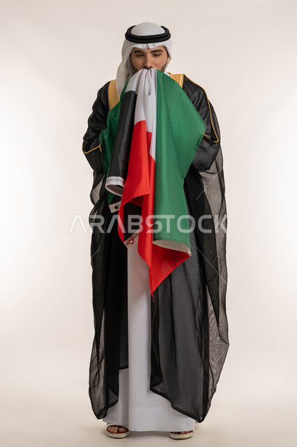 إظهار مشاعرحب الوطن والانتماء إليه, ارتداء الأزياء التقليدية في الأعياد والمناسبات الوطنية, بورتريه لشاب عربي خليجي كويتي يرتدي البشت والغترة يقوم بتقبيل العلم الكويتي بفخر واعتزاز, اليوم الوطني لدولة الكويت في 25 فبراير, صورة الجسم بالكامل, خلفية بيضاء