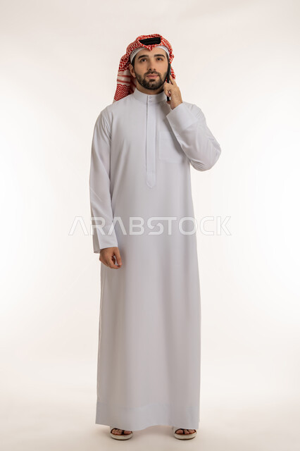 الدردشة والتواصل مع الأهل والأصدقاء عبر السوشال ميديا، اجراء مكالمة صوتية عبر الهاتف المحمول, بورتريه لشاب عربي خليجي سعودي يرتدي الثوب التقليدي والشماخ يمسك بيده الجوال, استخدام الاجهزة والتقنيات الحديثة والمتطورة، صورة الجسم بالطول الكامل, خلفية بيضاء