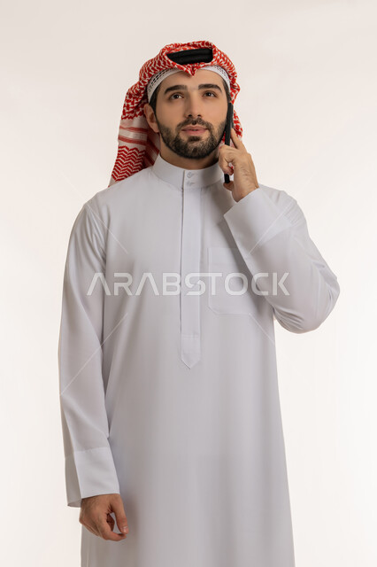 Using modern and advanced devices and technologies, looking at a place and trying to remember something specific, chatting and communicating with family and friends via social media, portrait of a young Saudi Arabian Gulf man wearing traditional thobe and shemagh making a voice call via mobile phone, white background