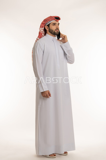 اجراء مكالمة صوتية عبر الهاتف المحمول, بورتريه لشاب عربي خليجي سعودي يرتدي الثوب التقليدي والشماخ يمسك بيده الجوال, استخدام الاجهزة والتقنيات الحديثة والمتطورة, الدردشة والتواصل مع الأهل والأصدقاء عبر السوشال ميديا، صورة الجسم بالطول الكامل, خلفية بيضاء