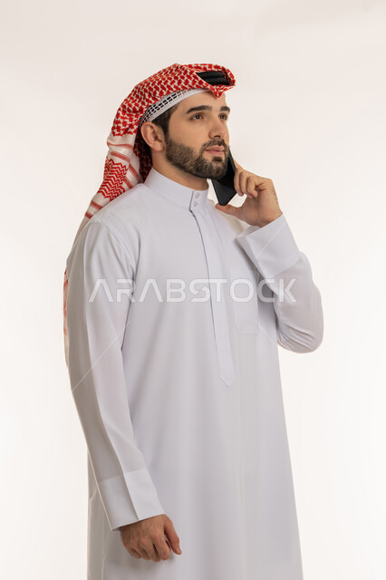 الدردشة والتواصل مع الأهل والأصدقاء عبر السوشال ميديا, اجراء مكالمة صوتية عبر الهاتف المحمول, بورتريه مقرب لشاب عربي خليجي سعودي يرتدي الثوب التقليدي والشماخ  يمسك بيده الجوال وينظر إلى شيء ما, ستخدام الاجهزة والتقنيات الحديثة والمتطورة، خلفية بيضاء
