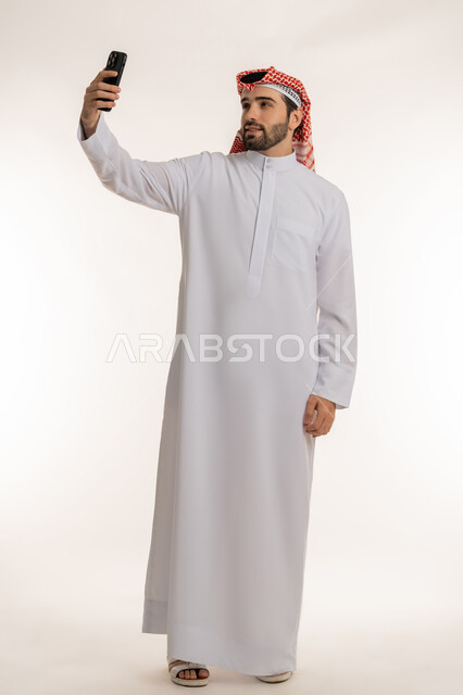 استخدام الاجهزة والتقنيات الحديثة والمتطورة, إجراء مكالمة فيديو عبر السوشال ميديا, بورتريه لشاب عربي خليجي سعودي يرتدي الثوب التقليدي والشماخ يمسك بيده الجوال, الدردشة والتواصل مع الأهل والأصدقاء عبر السوشال ميديا، صورة الجسم بالطول الكامل, خلفية بيضاء
