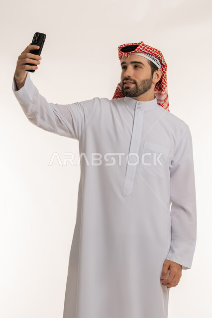 Chatting and communicating with family and friends via social media, using modern and advanced devices and technologies, making a video call via social media, portrait of a young Saudi Arabian Gulf man wearing traditional thobe and shemagh holding a mobile phone in his hand, white background