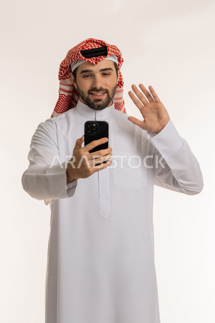 Using modern and advanced devices and technologies, making a video call via social media, portrait of a young Saudi Arabian Gulf man wearing traditional thobe and shemagh holding a mobile phone in his hand, chatting and communicating with family and friends via social media, white background