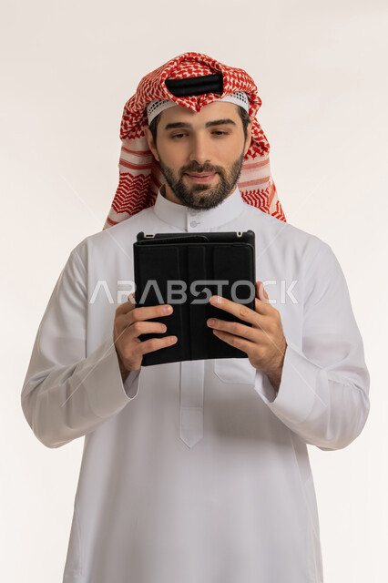 التواصل والدردشات مع الأهل والأصدقاء عبر السوشال ميديا، انجاز المهام عبر الأجهزة والتقنيات الحديثة والمتطورة, بورتريه مقرب لرجل عربي خليجي سعودي يرتدي الثوب التقليدي والشماخ يحمل بيده الجهاز اللوحي, خلفية بيضاء