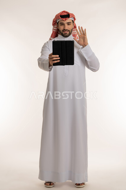 الدردشة والتواصل مع الأهل والأصدقاء عبر الأونلاين, إجراء مكالمة فيديو عبر السوشال ميديا, بورتريه لشاب عربي خليجي سعودي يرتدي الثوب التقليدي والشماخ يحمل بيده جهاز التابليت, استخدام الاجهزة والتقنيات الحديثة والمتطورة,  صورة الجسم بالطول الكامل, خلفية بيضاء