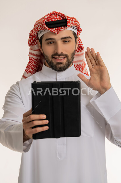 استخدام الاجهزة والتقنيات الحديثة والمتطورة, الدردشة والتواصل مع الأهل والأصدقاء عبر الأونلاين, إجراء مكالمة فيديو عبر السوشال ميديا, بورتريه لشاب عربي خليجي سعودي يرتدي الثوب التقليدي والشماخ يحمل بيده جهاز التابليت, خلفية بيضاء