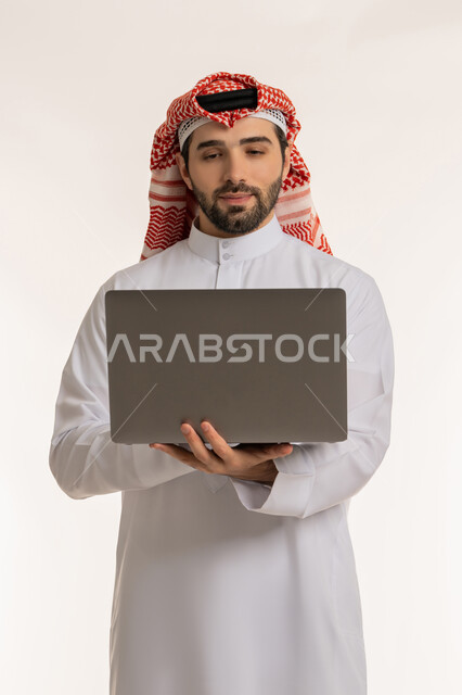 انجاز وادارة المهام عبر الأونلاين, التواصل والدردشة مع الأصدقاء والأهل عبر السوشال ميديا, مفهوم العمل عن بعد, دمج العمل مع التقنية والتكنولوجيا الحديثة والمتطورة,  بورتريه لشاب عربي خليجي سعودي يرتدي الثوب التقليدي والشماخ يستخدم الحاسب المحمول,  خلفية بيضاء