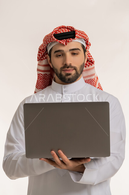 التواصل والدردشة مع الأصدقاء والأهل عبر السوشال ميديا, مفهوم العمل عن بعد, دمج العمل مع التقنية والتكنولوجيا الحديثة والمتطورة, انجاز وادارة المهام عبر الأونلاين, بورتريه لشاب عربي خليجي سعودي يرتدي الثوب التقليدي والشماخ يستخدم الحاسب المحمول,  خلفية بيضاء