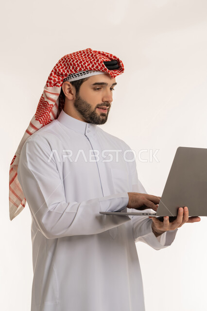 The concept of remote work, integrating work with modern and advanced technology and technology, accomplishing and managing tasks online, portrait of a young Saudi Arabian Gulf man wearing traditional thobe and shemagh using a laptop, communicating and chatting with friends and family via social media, white background