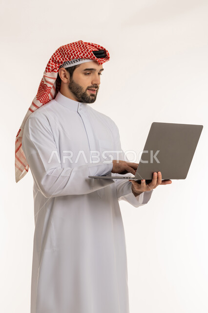 Integrating work with technology and modern and advanced technology, accomplishing and managing tasks online, the concept of remote work, a portrait of a young Saudi Gulf Arab man wearing traditional thobe and shemagh using a laptop, communicating and chatting with friends and family via social media, white background