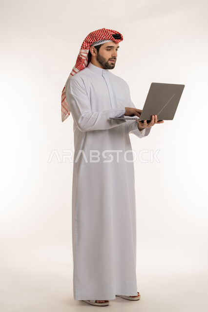 انجاز وادارة المهام عبر الأونلاين, مفهوم العمل عن بعد, بورتريه لشاب عربي خليجي سعودي يرتدي الثوب التقليدي والشماخ و يستخدم الحاسب المحمول, دمج العمل مع التقنية والتكنولوجيا الحديثة والمتطورة, صورة الجسم بالطول الكامل, خلفية بيضاء