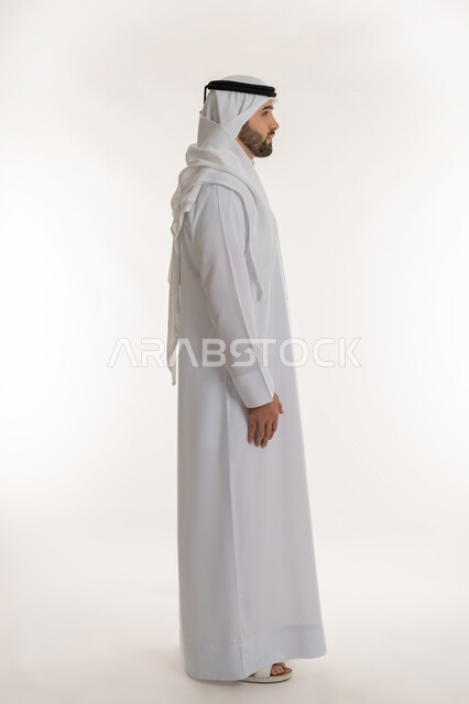 مفهوم الأناقة والرجولة, النظر الى مكان ما بايماءات الثقة بالنفس، الاهتمام بالمظهر الخارجي, بورتريه من الجانب لرجل عربي خليجي سعودي يرتدي الثوب التقليدي والغترة يقف باستقامة واعتدال, الجسم بالطول الكامل, خلفية بيضاء