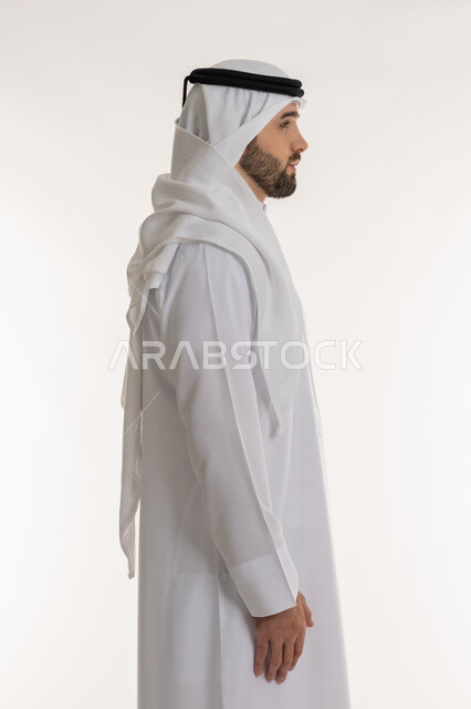 Looking at a place with gestures of self-confidence, interest in appearance, concept of elegance and masculinity, side portrait of a Saudi Arabian Gulf man wearing traditional thobe and ghutra standing straight and upright, white background