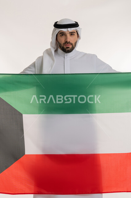 الحب والانتماء للوطن، النظر إلى الكاميرا بايماءات الاعتزاز والفخر بإنجازات البلاد، اليوم الوطني الكويتي في 25 فراير، بورتريه لشاب عربي خليجي كويتي يرتدي الثوب التقليدي والغترة يحمل علم دولة الكويت، خلفية بيضاء