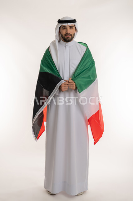 مناسبات وأعياد وطنية, اظهار مشاعر الفخر بالانتماء للوطن, بورتريه لشاب عربي خليجي كويتي يرتدي الثوب التقليدي والغترة يضع على ظهره علم دولة الكويت باعتزاز, استذكار اليوم الوطني الكويتي في 25 فبراير, صورة الجسم بالطول الكامل, خلفية بيضاء