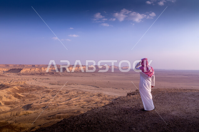 التشكيلات والتكوينات الصخرية والمناظر الطبيعية في الصحراء، حافة نهاية العالم الجبلية في مدينة الرياض في المملكة العربية السعودية ، المعالم والأماكن السياحية الطبيعية المشهورة ، صورة من الخلف لرجل عربي سعودي يرتدي الثوب والشماغ يقف باستقامة