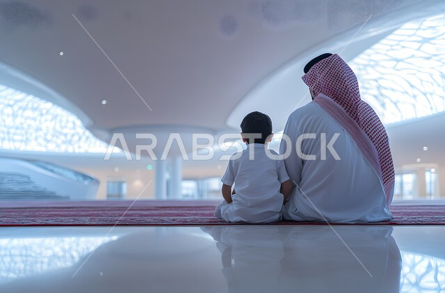  أداء فريضة الصلاة، الاجتهاد في العبادة والطاعة في شهر رمضان المبارك، التوبة والرجوع إلى الله، مفهوم الدعاء والتضرع والترجي، صورة من الخلف لرجل عربي خليجي سعودي يرتدي الثوب التقليدي يجلس مع ابنه داخل المسجد