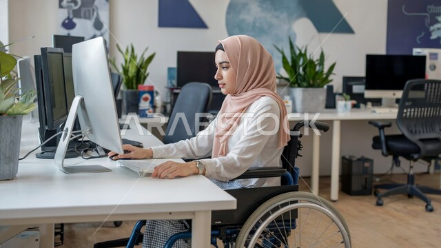 Women's jobs and professions, completing work tasks remotely, using modern and advanced technologies, a disabled Saudi Arabian Gulf woman wearing casual clothes sitting in a wheelchair working on a laptop, following up and developing businesses