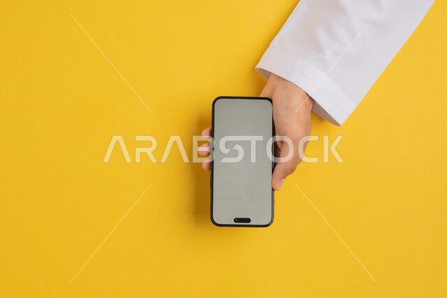 عرض شاشة رمادية فارغة، بورتريه علوي عامودي ليدين رجل عربي خليجي إماراتي يقوم بحمل الهاتف المحمول، شاب سعودي يقوم بمتابعة الدروس التعليمية أونلاين، البيع والشراء عبر الانترنت، الترويج والتسويق الالكتروني، تصفح مواقع التواصل الاجتماعي، خلفية صفراء