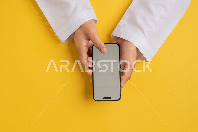 Integrating technology and modern advanced techniques in work, vertical portrait of an Arab Gulf Emirati man holding a mobile phone, a young Saudi man browsing social media, doing e-marketing, clicking on a blank gray screen, yellow background