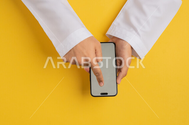النقر على شاشة رمادية فارغة، دمج التكنولوجيا والتقنيات الحديثة المتطورة بالعمل، بورتريه علوي عامودي ليدين رجل عربي خليجي إماراتي يحمل الهاتف المحمول، شاب سعودي يقوم بتصفح مواقع التواصل الاجتماعي، القيام بعمليات التسويق الالكتروني، خلفية صفراء