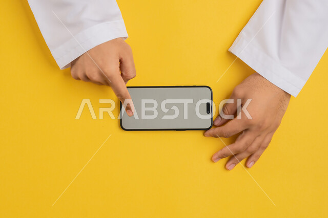 القيام بعمليات التسويق الالكتروني، النقر على شاشة رمادية فارغة، دمج التكنولوجيا والتقنيات الحديثة المتطورة بالعمل، بورتريه علوي عامودي ليدين رجل عربي خليجي إماراتي يحمل الهاتف المحمول، شاب سعودي يقوم بتصفح مواقع التواصل الاجتماعي، خلفية صفراء