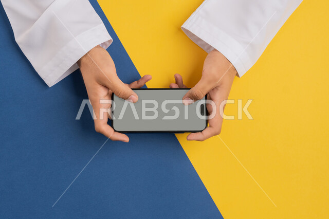 Browsing social media, blank gray screen display, vertical top portrait of an Arab Gulf Emirati man holding a mobile phone, a Saudi young man following educational lessons online, buying and selling online, e-marketing and promotion, colorful background