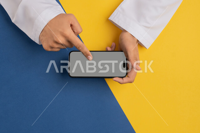 Promotion and e-marketing, browsing social media, displaying a blank gray screen, vertical top portrait of an Arab Gulf Emirati man holding a mobile phone, a young Saudi man following educational lessons online, buying and selling online, colorful background