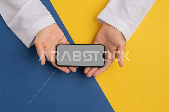 البيع والشراء عبر الانترنت، الترويج والتسويق الالكتروني، تصفح مواقع التواصل الاجتماعي، عرض شاشة رمادية فارغة، بورتريه علوي عامودي ليدين رجل عربي خليجي إماراتي يقوم بحمل الهاتف المحمول، شاب سعودي يقوم بمتابعة الدروس التعليمية أونلاين، خلفية ملونة