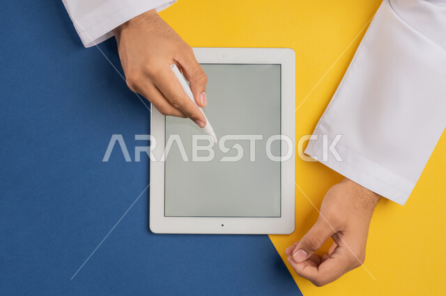 عرض شاشة رمادية فارغة، بورتريه علوي عامودي ليدين رجل عربي خليجي إماراتي يقوم بإنجاز المهام المطلوبة عن بعد، شاب سعودي يستخدم جهاز الآيباد، رسم الصور والتصاميم الالكترونية، استعمال قلم التابلت لتدوين ملاحظات خاصة بالعمل، متابعة الدروس التعليمية، خلفية ملونة